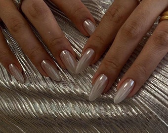 May include: Close-up of hands with long, almond-shaped nails painted with a pearlescent white polish. The nails have a subtle shimmer and are accented with gold rings.