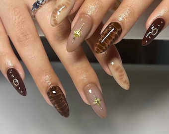 May include: Close-up of a hand with long, almond-shaped nails painted in a brown and beige color scheme. Some nails feature a snakeskin pattern, while others have gold star accents.