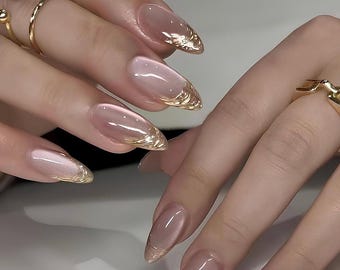 May include: Close-up of a hand with long, almond-shaped nails painted with a sheer nude polish and accented with a gold foil design at the tips.