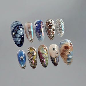 May include: A set of ten press-on nails featuring diverse designs including dark blue butterfly wing patterns, iridescent finishes, silver metallic 3D liquid accents, star motifs, and swirling abstract colour blends with small silver spheres.
