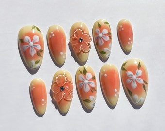 Op de afbeelding: Set van tien kunstnagels met een bloemenontwerp. Elke nagel heeft een gele tot perzikkleurige gradiënt, met witte bloemen en groene bladeren. Sommige nagels hebben 3D perzikbloemen met een strass steentje in het midden.