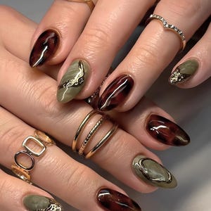 Può includere: Primo piano di una mano con unghie a mandorla dipinte con uno smalto tartaruga e verde. Alcune unghie hanno accenti dorati. La mano indossa diversi anelli dorati.