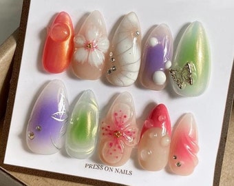 May include: A set of ten press-on nails with various designs. The nails are in different colors, including pink, purple, green, and white. Some nails have floral designs, while others have butterfly or geometric designs. The nails are arranged in two rows on a white card with the text "PRESS ON NAILS".