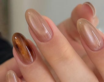 Puede incluir: Primer plano de una mano con uñas largas y ovaladas. Las uñas están pintadas en una variedad de tonos neutros, incluyendo marrón claro y un marrón más oscuro y veteado. Las uñas tienen un acabado brillante, reflejando la luz.