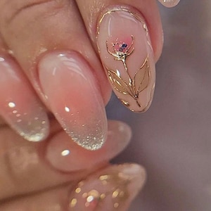 Könnte beinhalten: Nahaufnahme einer Hand mit langen, ovalen Nägeln, die mit einem hellrosa und weißen Ombre-Effekt lackiert sind. Die Nägel sind mit goldenen Blatt-Akzenten und einem zarten Blumenmuster verziert.