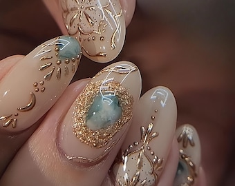 May include: Close-up of a hand with intricately designed nail art. The nails are a light beige color, adorned with gold embellishments and accents. Some nails feature a gold glitter border around a blue-green stone.