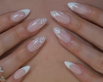 May include: Close-up of a hand with almond-shaped nails painted with a white tip and a delicate white floral design.