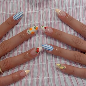 Puede incluir: Uñas largas en forma de almendra con varios diseños. Algunas uñas presentan rayas azules y blancas, otras ilustraciones de frutas como naranjas, cerezas y fresas. Las uñas están pintadas sobre un fondo a rayas rosas y blancas.