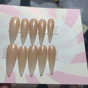 May include: Ten nude-colored, long, stiletto-shaped fake nails with a gradient effect, going from a light nude to a darker nude color. The nails are arranged in two rows of five on a white and pink background with a swirl pattern.