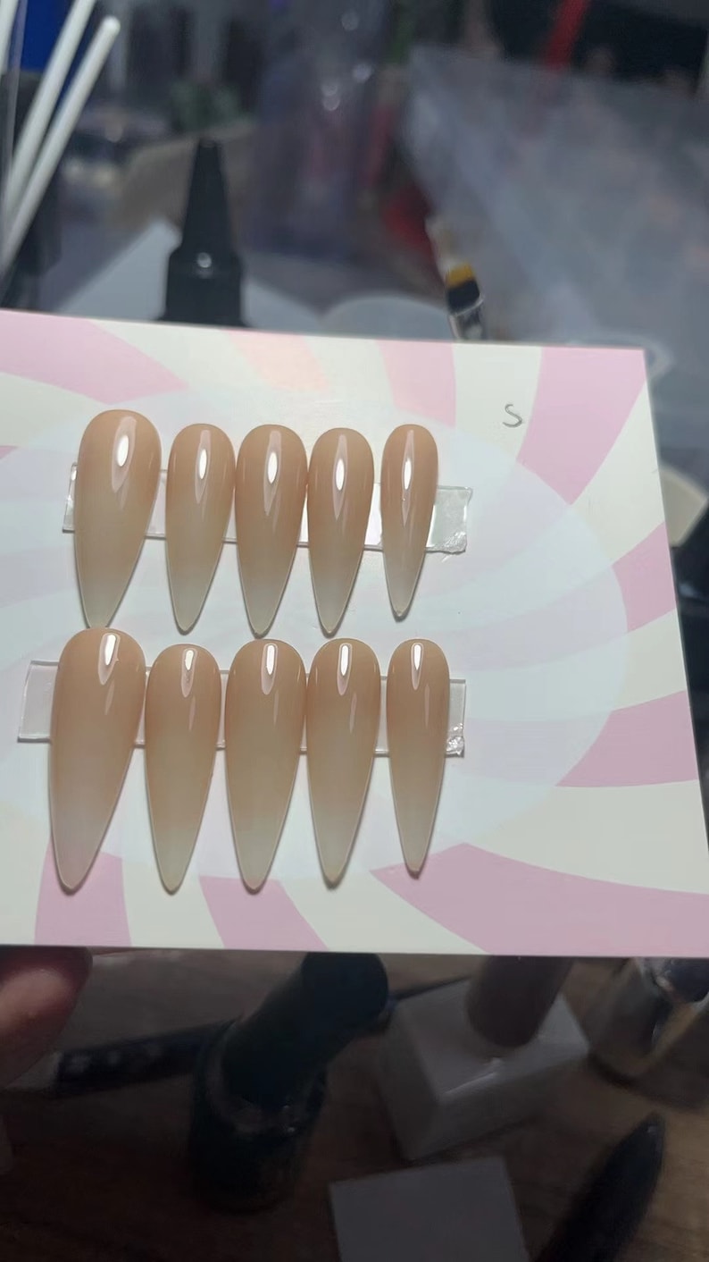 May include: Ten nude-colored, long, stiletto-shaped fake nails with a gradient effect, going from a light nude to a darker nude color. The nails are arranged in two rows of five on a white and pink background with a swirl pattern.