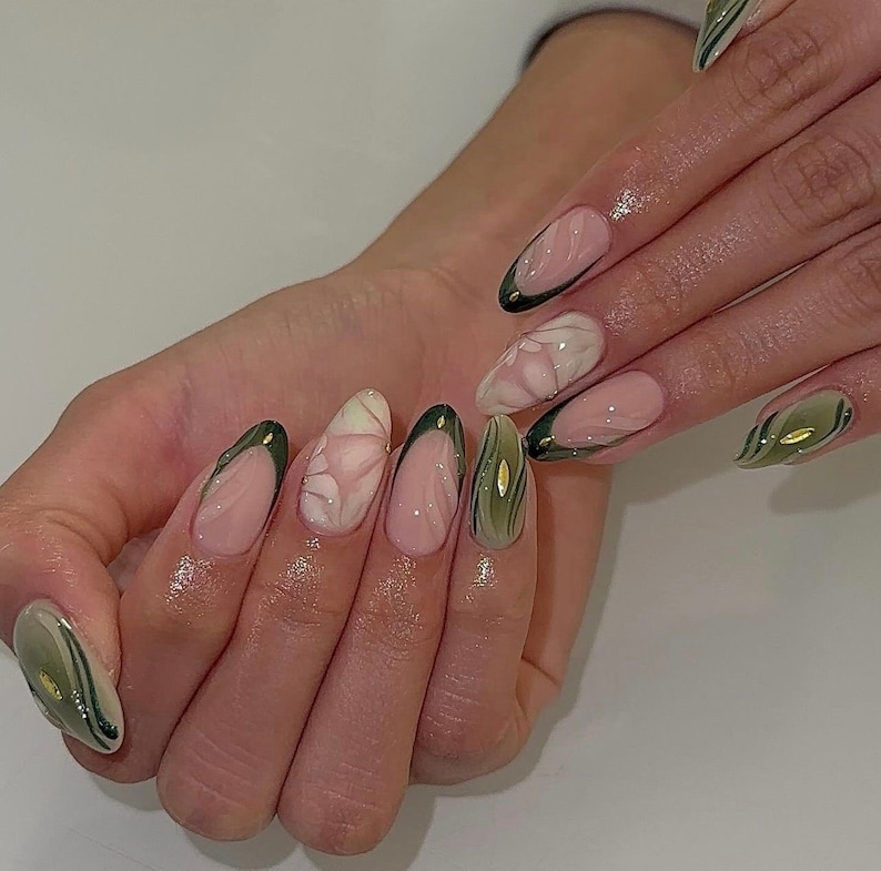 May include: Close-up of a hand with almond-shaped nails painted with a light pink base and green and gold abstract designs.