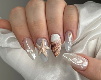 May include: Close-up of a hand with several nails painted in a light gray color. Each nail has a different design, including a seashell, a starfish, and a small pearl.