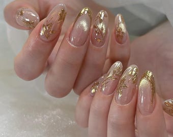 May include: Close-up of nude-colored nails with a gold leaf design. The nails are long and almond-shaped.