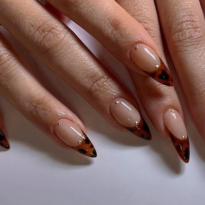 May include: Close-up of a hand with long, almond-shaped nails painted with a nude polish. The tips of the nails are accented with a brown and black tortoise shell design and small gold dots.