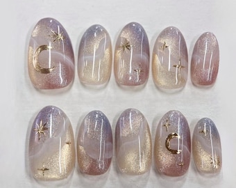 Puede incluir: Un conjunto de diez uñas postizas ovaladas. Cada uña presenta un diseño degradado con colores que van del dorado al morado y rosa. Se aplican lunas crecientes y estrellas doradas en algunas uñas.