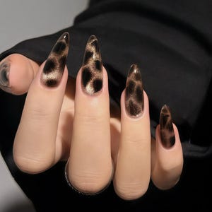 May include: A hand displaying long, pointed fingernails with a leopard print design. The nails are a clear base with brown and black spots, and a glittery gold shimmer. The nails are a fashionable accessory.