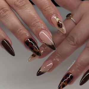 May include: Close-up of a hand with long, almond-shaped nails painted with a brown and gold tortoiseshell design. Some nails have a thin gold line along the edge.