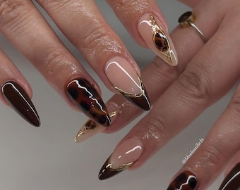 May include: Close-up of a hand with long, almond-shaped nails painted with a brown and gold tortoiseshell design. Some nails have a thin gold line along the edge.