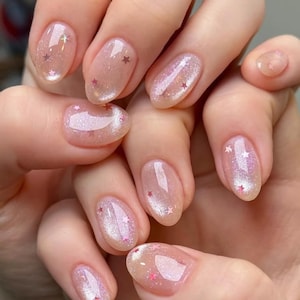 May include: Close-up of a hand with oval-shaped nails painted with a sheer, light pink nail polish with iridescent glitter and small pink stars.