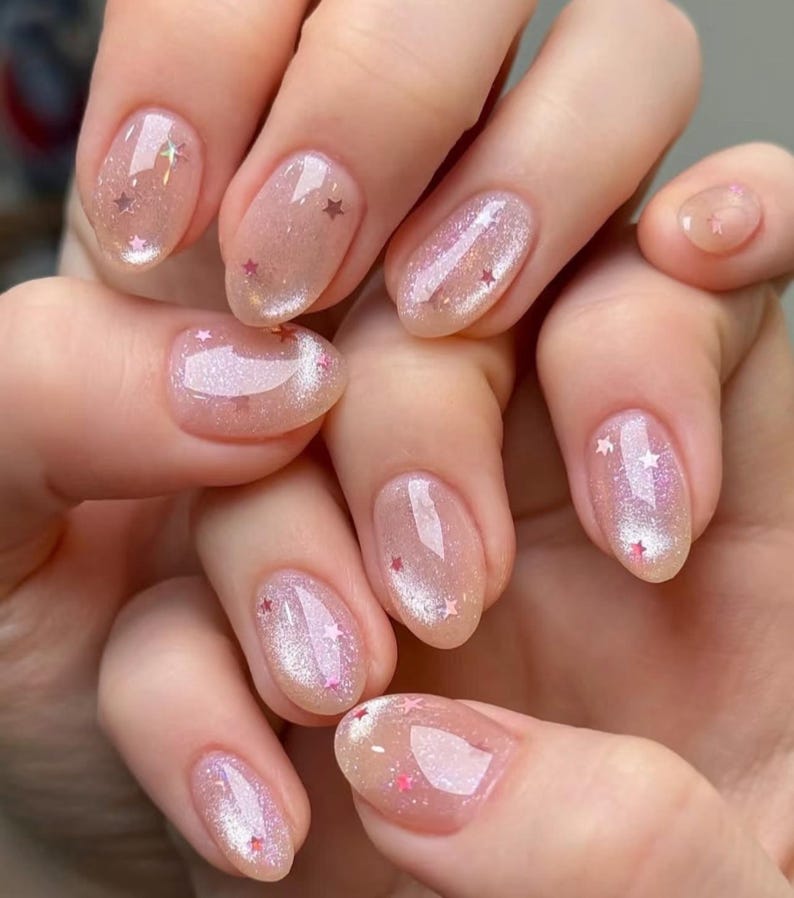 May include: Close-up of a hand with oval-shaped nails painted with a sheer, light pink nail polish with iridescent glitter and small pink stars.