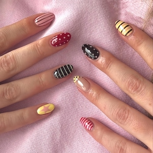 Peut inclure: Gros plan sur une main avec différents motifs de nail art. Les ongles présentent des motifs tels que des rayures, des pois et une étoile. Les couleurs incluent le rose, le rouge, le noir, le jaune et le nude. Le fond est un tissu rose clair.