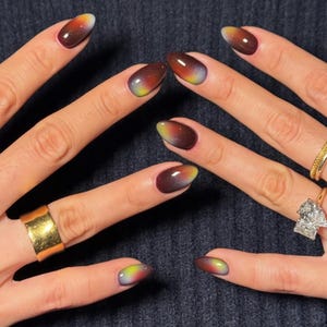 May include: Oval-shaped nails with a gradient design. The nails transition from a deep brown at the base to yellow, orange, and white at the tips. The hands also feature gold rings, including a wide band and a diamond-studded ring.