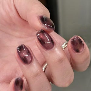 May include: Close-up of a hand with oval-shaped nails painted with a dark, translucent plum colour. The nails have a glossy finish, and the colour appears to have a subtle, smoky effect. A silver ring is visible on one finger.