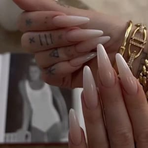 May include: Close-up of long, almond-shaped nails with a white ombre design. The nails are painted with a light pink base that fades to white at the tips. The nails are long and slender, and the ombre effect is subtle and elegant.