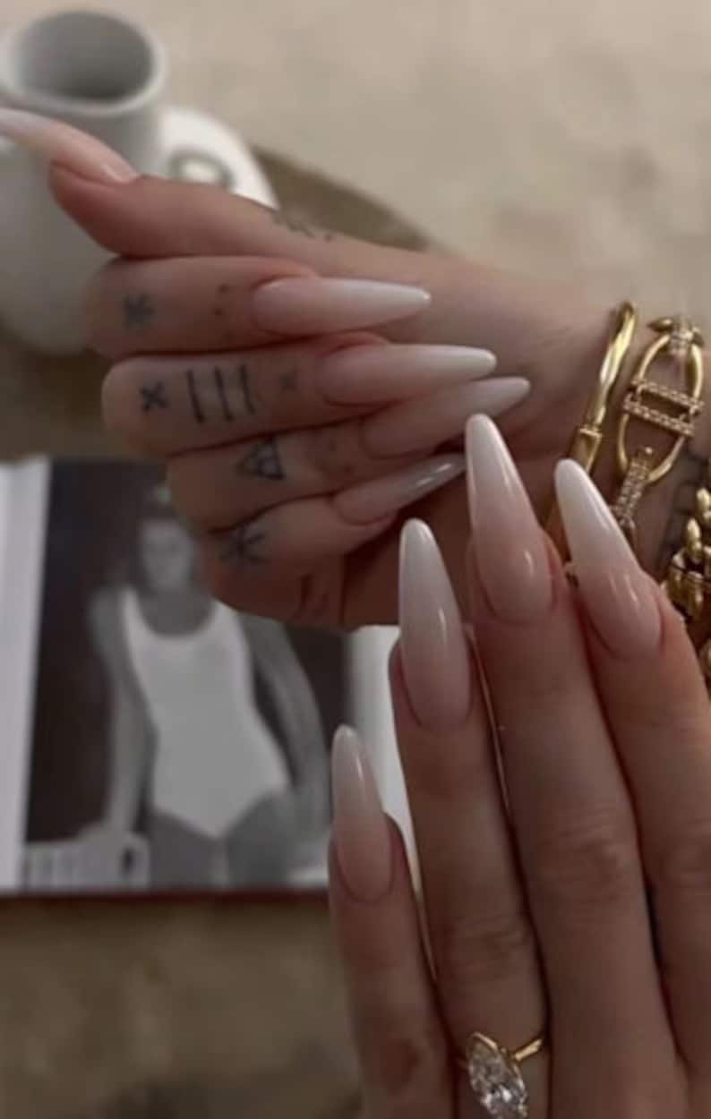 May include: Close-up of long, almond-shaped nails with a white ombre design. The nails are painted with a light pink base that fades to white at the tips. The nails are long and slender, and the ombre effect is subtle and elegant.