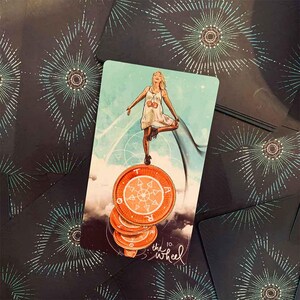 May include: A tarot card featuring a woman balancing on a stack of orange coins, with the text "the wheel". The card has a light blue background with a woman in a white dress. The card is surrounded by other cards with a dark background and white starburst design.