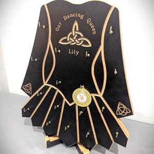May include: Black wooden medal display shaped like a dress, with gold trim and hooks. The text "Our Dancing Queen Lily" is engraved on the top. A gold medal hangs from the center. A decorative piece for displaying medals.