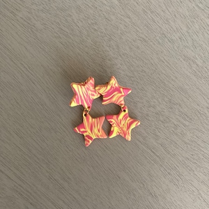 May include: Four star-shaped earrings with a marbled design in yellow and pink. The earrings are connected with gold-coloured jump rings. The earrings are a fun and colourful accessory.