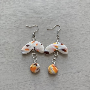 Puede incluir: Un par de pendientes colgantes con un diseño marmoleado blanco, naranja y marrón. Los pendientes tienen una pieza superior curva y una pieza inferior redonda conectadas por una cadena plateada.