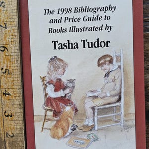 May include: A book cover with a red background and a white title that reads "The 1998 Bibliography and Price Guide to Books Illustrated by Tasha Tudor". The cover features an illustration of two children sitting in chairs with a dog and a cat. The text "Compiled by Gary J. Overmann" is at the bottom of the cover.