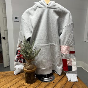 May include: A gray hooded sweatshirt with red, white, and black patchwork fabric on the sleeves. The sweatshirt is on a wooden table with a small potted plant in front of it.