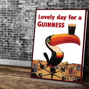 K&ouml;nnte beinhalten: Ein gerahmtes Guinness-Poster mit einem Tukan, der ein Pint Guinness h&auml;lt. Der Text "Lovely day for a Guinness" ist rot. Der Tukan hat einen gro&szlig;en orange-roten Schnabel. Der Hintergrund zeigt eine Stadt und einen Kompass.