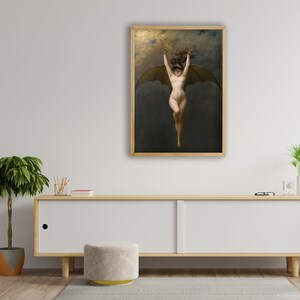 May include: A framed artwork depicts a nude figure with bat-like wings against a dark, cloudy background. The figure is suspended mid-air with arms raised. The artwork is in a wooden frame and hangs on a white wall above a wooden cabinet.