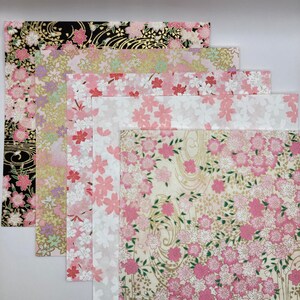 Könnte beinhalten: Ein Set aus sechs gemusterten Papieren mit rosa und weißen Blumenmustern und goldenen Akzenten. Die Papiere sind in einem Stapel angeordnet, wobei das größte Papier unten und das kleinste Papier oben liegt.