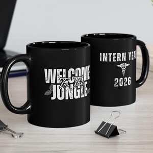 May include: Two black ceramic mugs featuring white printed text. One mug displays the words "WELCOME TO THE JUNGLE" with leaf accents. The other mug features the text "INTERN YEAR" above a medical caduceus symbol and the year "2026" below it.