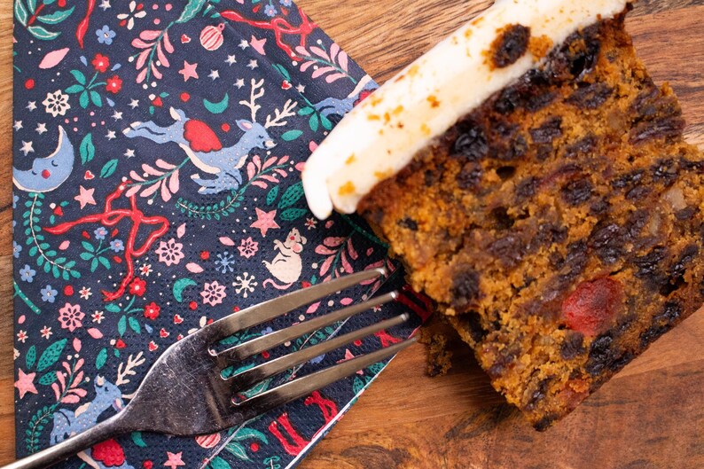 May include: A slice of fruitcake with white icing, alongside a festive napkin and a fork. The napkin has a navy blue background with a winter holiday design of reindeer, stars, and floral elements. The cake is on a wooden surface.
