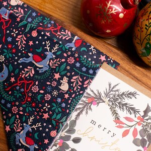 May include: A navy blue Christmas napkin featuring reindeer, stars, and festive ornaments. A greeting card with a wreath design and the words "merry christmas" is next to the napkin. Two decorative baubles are also in the image.