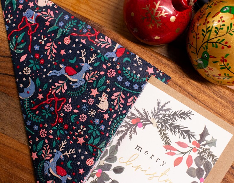 May include: A navy blue Christmas napkin featuring reindeer, stars, and festive ornaments. A greeting card with a wreath design and the words "merry christmas" is next to the napkin. Two decorative baubles are also in the image.
