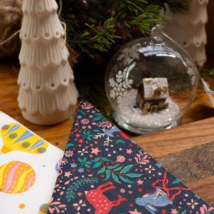 May include: A Christmas-themed arrangement with a white ceramic tree, a glass bauble containing a miniature house, and patterned napkins. The napkins showcase festive designs with reindeer and floral motifs.