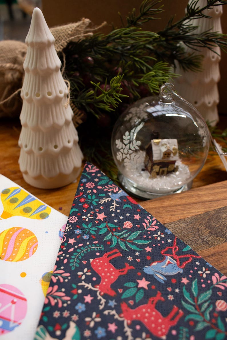 May include: A Christmas-themed arrangement with a white ceramic tree, a glass bauble containing a miniature house, and patterned napkins. The napkins showcase festive designs with reindeer and floral motifs.