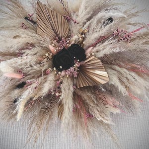 May include: A decorative floral arrangement featuring fluffy beige pampas grass, dried palm leaves, and small pink sprigs. Two prominent black roses are positioned in the centre, surrounded by a mix of textured dried grasses and foliage.