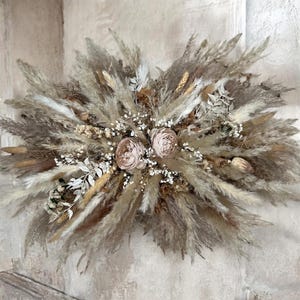 May include: A large dried floral arrangement with pampas grass, wheat, and white accents. Two large, pale pink roses are at the centre. The neutral tones create a natural, bohemian aesthetic, ideal for home decor.