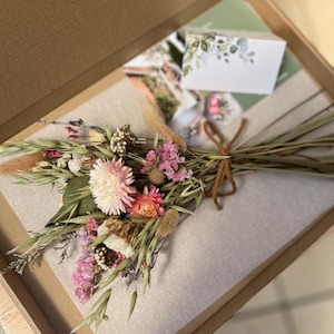 May include: A small bouquet of dried flowers and grasses tied with brown twine, featuring pink, white, and green tones. The bouquet rests on a piece of light grey paper inside a brown cardboard box, accompanied by a small white card with floral print.