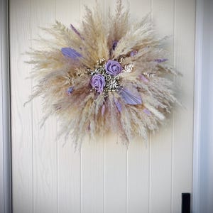 May include: A circular decorative wreath composed of fluffy beige pampas grass, dried foliage, and small white filler flowers. Two prominent lavender-coloured roses are positioned in the centre, accented by additional purple-dyed dried stems and leaves.