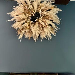 May include: A circular wall wreath composed of fluffy beige pampas grass, dried palm leaves, small white baby's breath flowers, and two prominent black roses at the centre. The arrangement features a mix of natural textures and dark floral accents.
