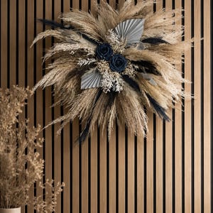 May include: A circular wall wreath composed of beige pampas grass, black plumes, and two dark navy blue roses. The arrangement features two grey fan-shaped palm leaves, white dried filler flowers, and delicate sprigs of white foliage.